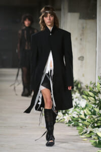 Model walking a runway in an oversized black coat with white layered undergarments and tall boots; headband adornment visible.