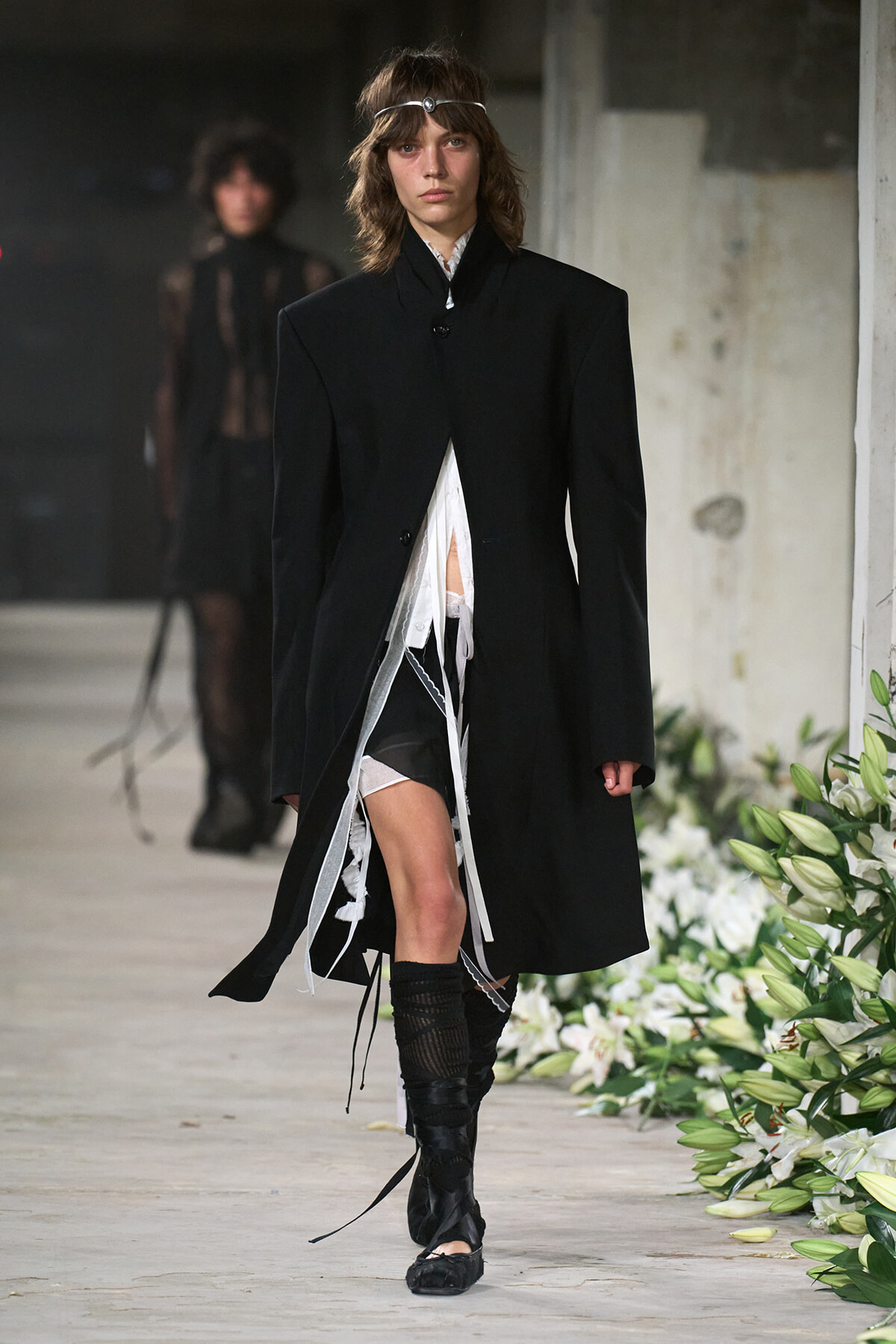 Model walking a runway in an oversized black coat with white layered undergarments and tall boots; headband adornment visible.