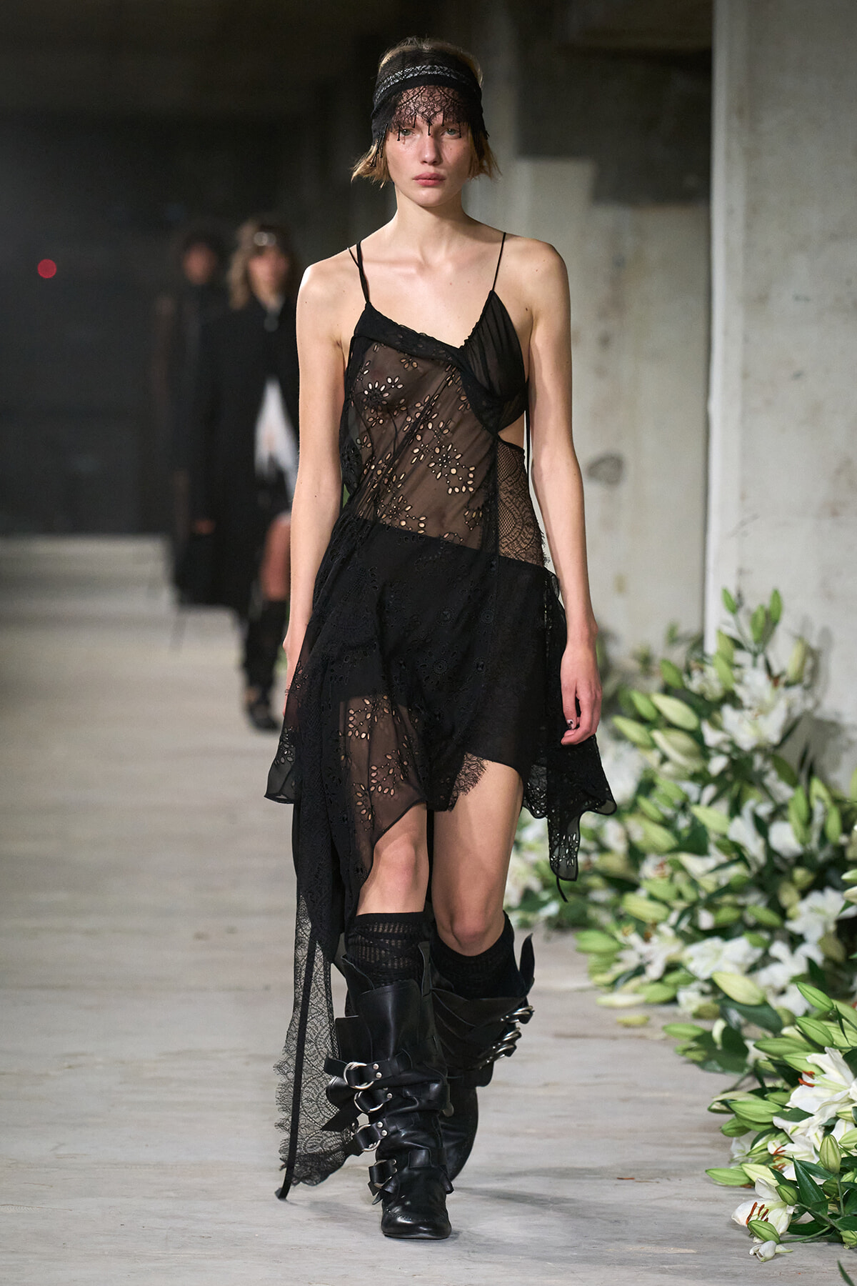 Model walking the runway in a sheer black lace dress with floral embroidery, layered asymmetrical skirt, lace headband, and buckled black boots.