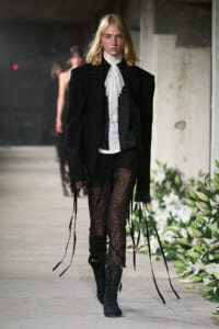 Model walking a runway in a black oversized blazer, white lace necktie, sheer lace pants, and black boots against an industrial backdrop with floral accents on the side.