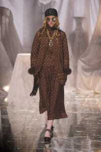 Model on a fashion runway wearing a brown patterned dress with fur cuffs, chunky gold chain, and round sunglasses.