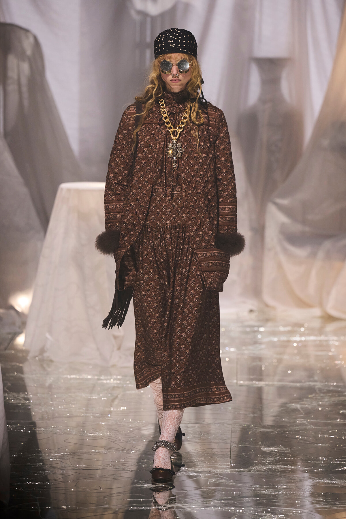 Model on a fashion runway wearing a brown patterned dress with fur cuffs, chunky gold chain, and round sunglasses.