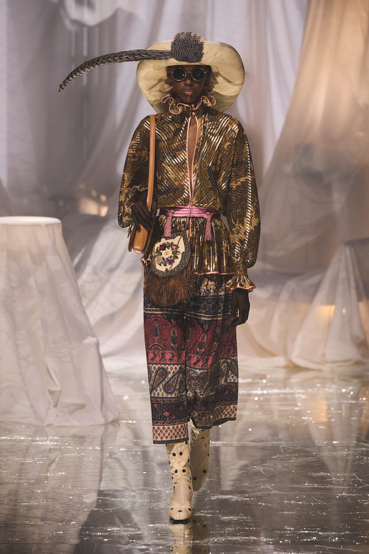 Model walks the runway in a metallic gold blouse with pink sash, a patterned skirt, oversized straw hat with a feather, round sunglasses, and cream boots, carrying a tan bag.
