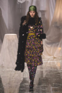Model on a runway wearing a black fur coat over a purple-yellow floral dress with a green satin headscarf.