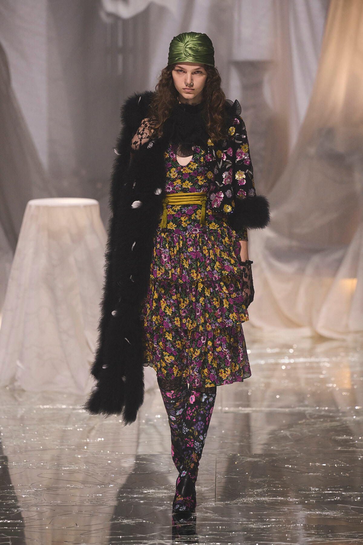 Model on a runway wearing a black fur coat over a purple-yellow floral dress with a green satin headscarf.