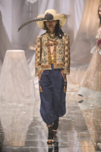 Model walking the runway in a wide-brimmed beige hat with a black feather, a beaded-embroidered jacket, and blue harem pants with tassel accents.