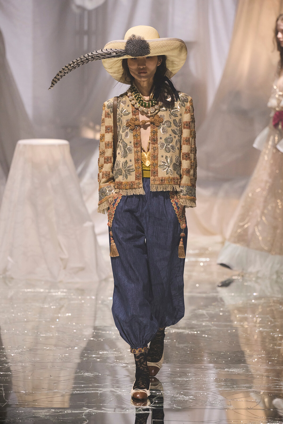 Model walking the runway in a wide-brimmed beige hat with a black feather, a beaded-embroidered jacket, and blue harem pants with tassel accents.