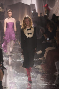 Model in a black long-sleeve gown with white bib and two bows walks the runway; red knee-high boots with sparkling accents visible. Audience watches on both sides.
