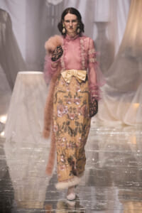 Model on a runway wearing a pink sheer blouse with a gold floral-embroidered skirt, pale yellow bow at the waist, fur stole, and black lace gloves.
