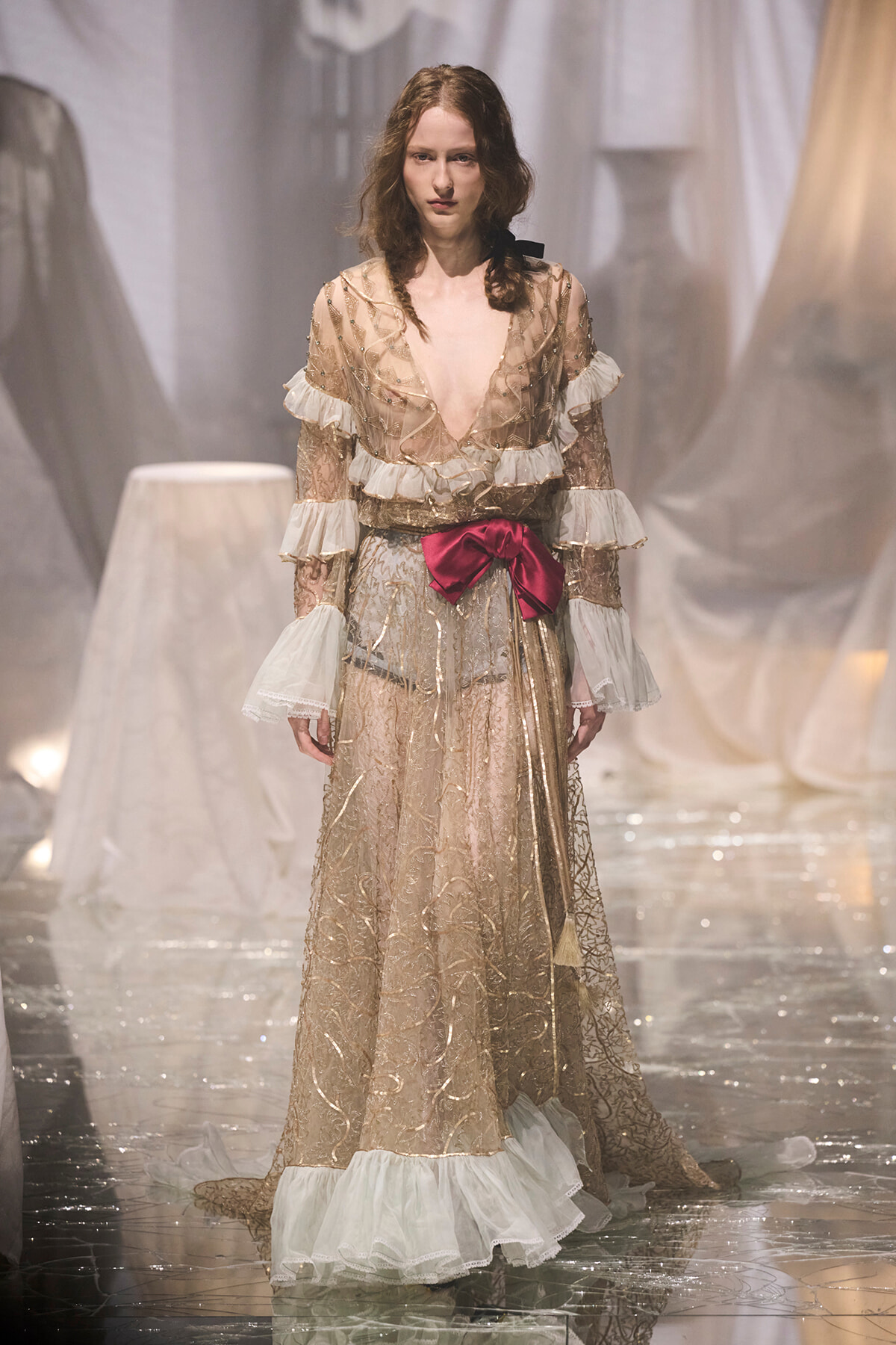 Model walking a fashion runway in a gold lace gown with sheer sleeves, white ruffles, and a pink bow at the waist.