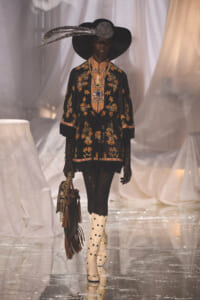 Model walking a fashion runway in a black floral-embroidered tunic, wide-brim hat with feather, lace gloves, cream polka-dot boots, and a fringed bag.