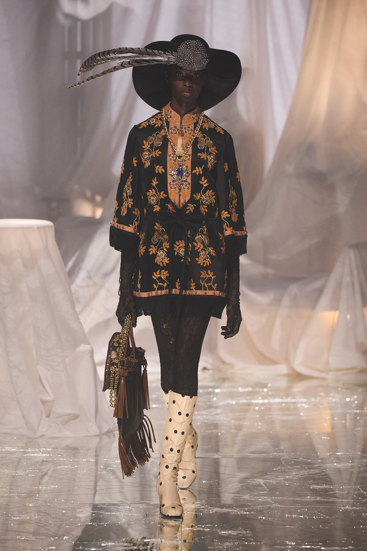 Model walking a fashion runway in a black floral-embroidered tunic, wide-brim hat with feather, lace gloves, cream polka-dot boots, and a fringed bag.