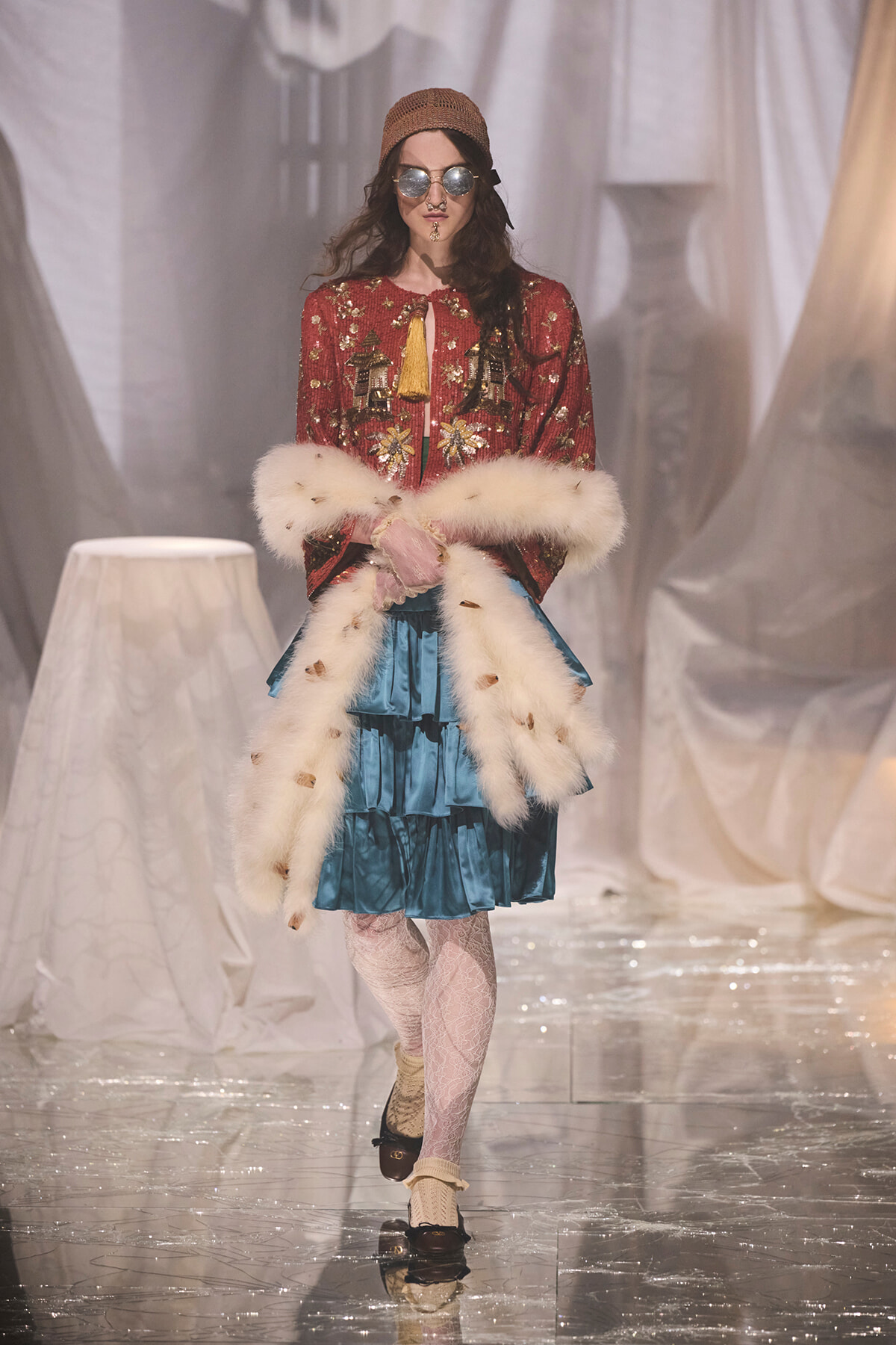Model walking runway in a red sequined jacket, fur stole, and teal ruffled skirt with a brown knit cap and round sunglasses.