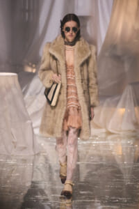 Model walking a runway in a beige fur coat over a pink lace dress, layered pearls, round sunglasses, and a black-and-cream handbag.