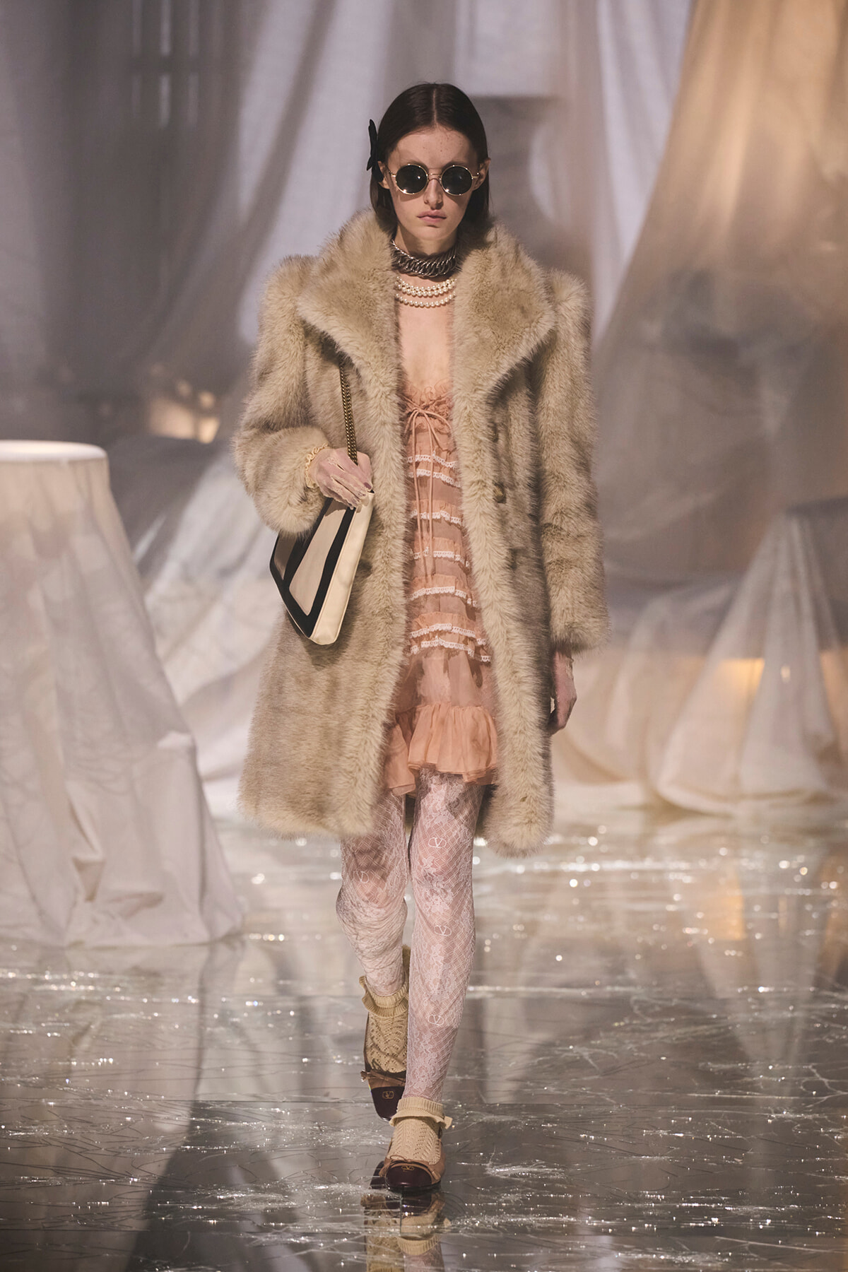 Model walking a runway in a beige fur coat over a pink lace dress, layered pearls, round sunglasses, and a black-and-cream handbag.
