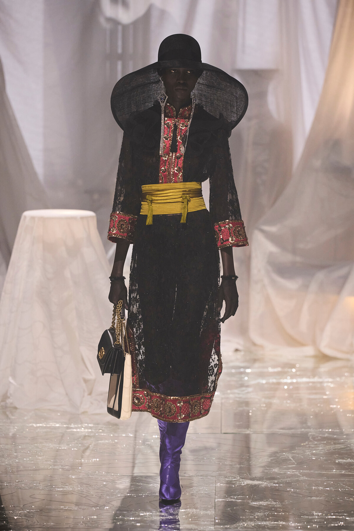 Model on runway in a black lace dress with red embroidery, wide yellow sash belt, oversized hat, purple boots, and a black-and-cream handbag.