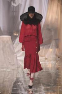 Model on a runway wearing a red, long-sleeved dress with a black ruffled collar, wide-brim hat, and white lace tights. A glossy runway gleams beneath her boots with gold buttons on the skirt edge.