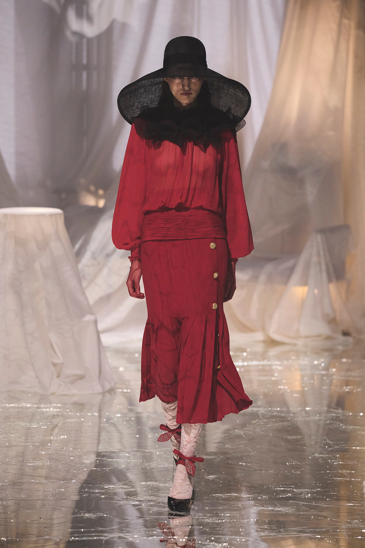 Model on a runway wearing a red, long-sleeved dress with a black ruffled collar, wide-brim hat, and white lace tights. A glossy runway gleams beneath her boots with gold buttons on the skirt edge.