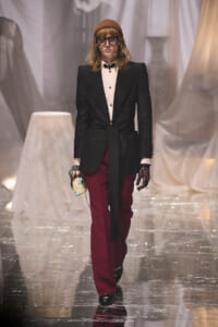 Model walking a runway in a black blazer with a belt, white shirt, burgundy pants, brown beanie, and chunky gloves, holding a small bag.