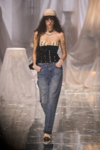 Model walking runway in a beige strapless top, denim jeans, hat, and pearl necklace with a small bag over the shoulder.