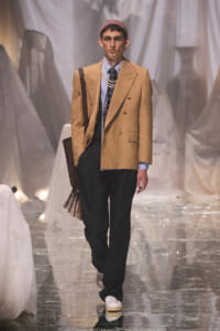 Male model on a runway wearing a camel double-breasted blazer, light blue shirt, patterned tie, layered pearls, beanie, black trousers, white sneakers, and a fringed brown bag.