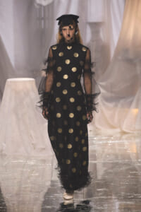 Model walks the runway wearing a black floor-length dress with gold polka dots, sheer sleeves, veil, and a black cap with netting.