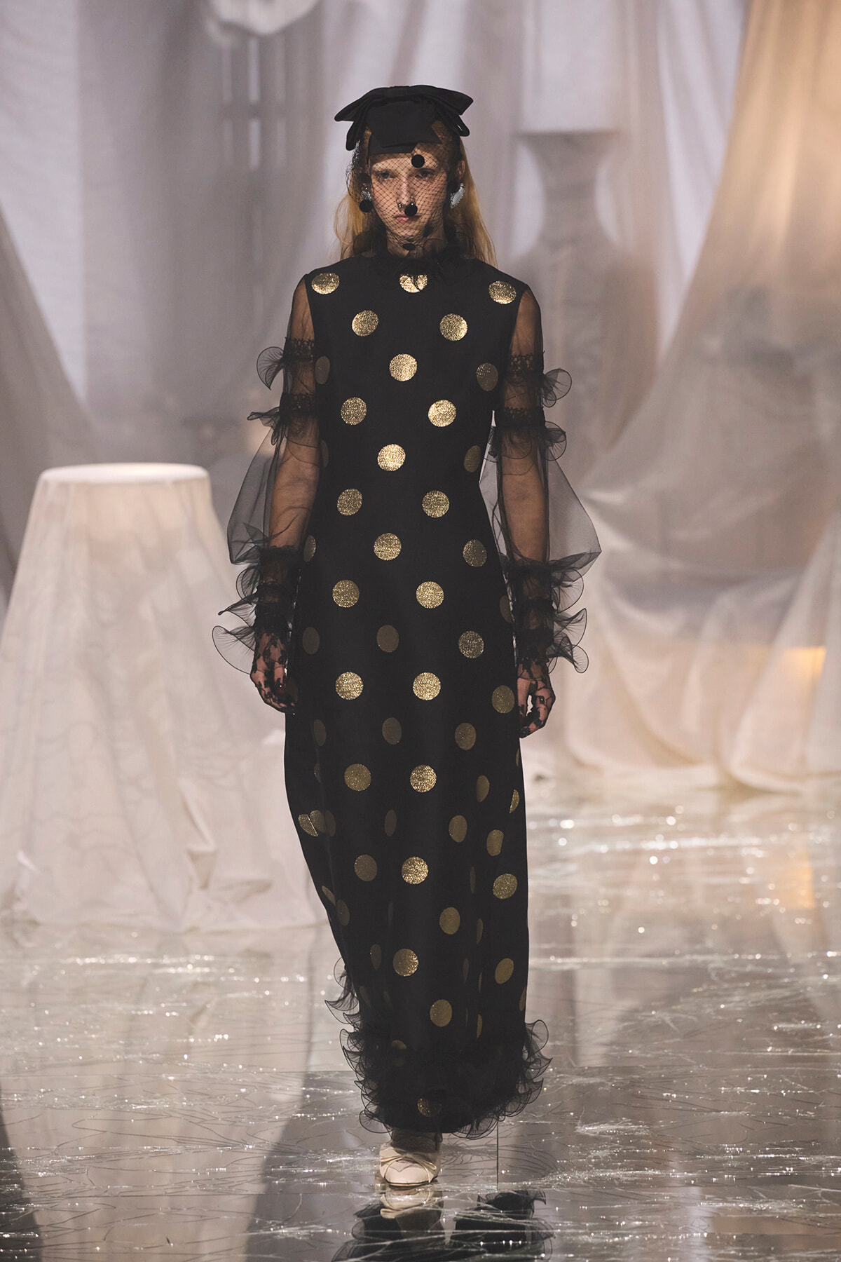 Model walks the runway wearing a black floor-length dress with gold polka dots, sheer sleeves, veil, and a black cap with netting.