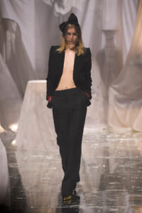 Model walking runway in an oversized black suit with no top beneath, black blazer open, matching trousers, red cuffs, and a large black bow headpiece with a sheer veil; gold shoes glide on a glossy runway with white fabric backdrop.