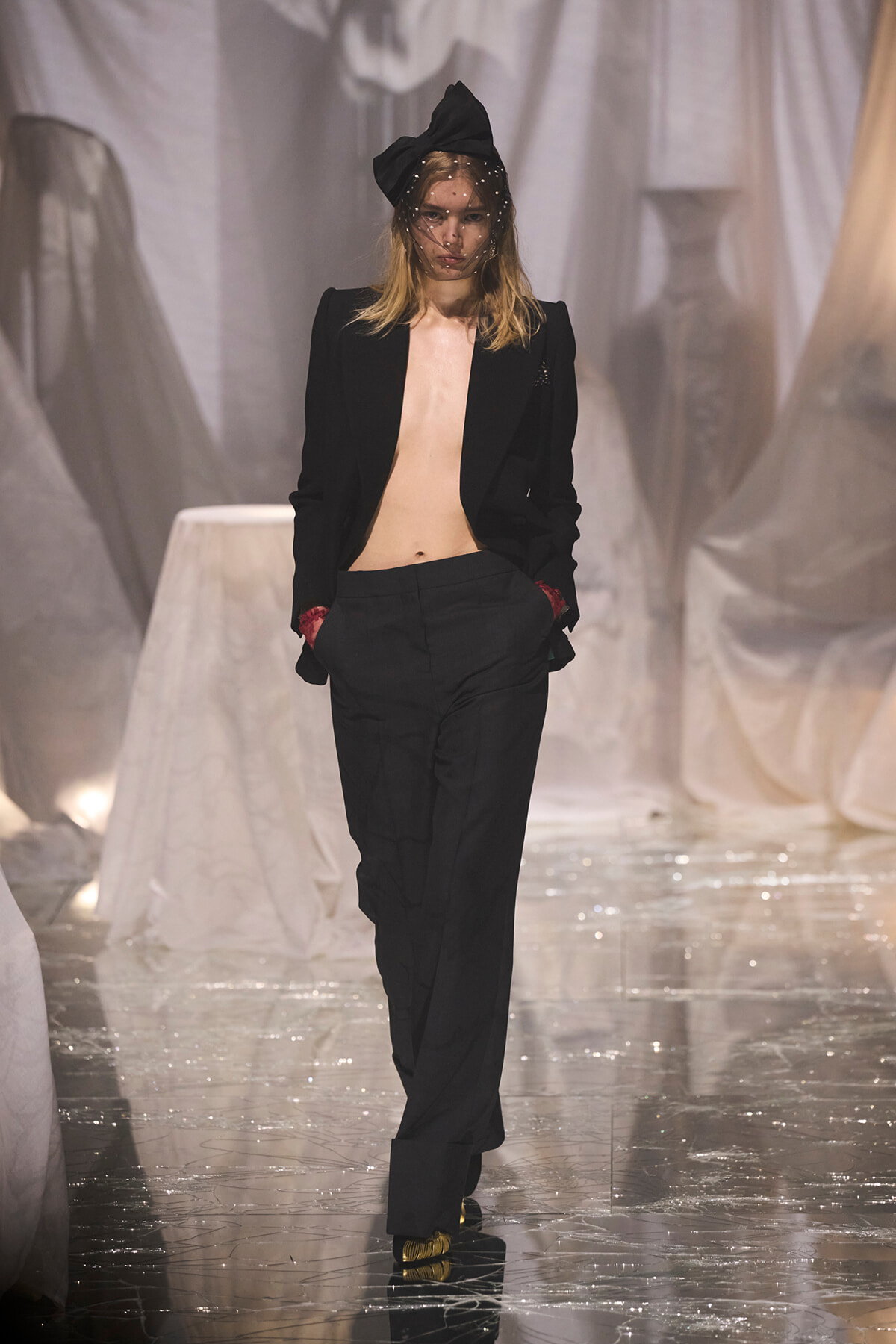 Model walking runway in an oversized black suit with no top beneath, black blazer open, matching trousers, red cuffs, and a large black bow headpiece with a sheer veil; gold shoes glide on a glossy runway with white fabric backdrop.