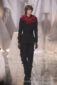 Model walks a runway in an all-black outfit with a large red ruffled collar, black lace gloves, and a wrapped waist belt. White draped backdrop in the background.