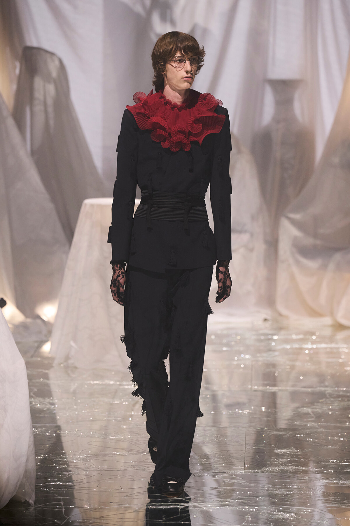 Model walks a runway in an all-black outfit with a large red ruffled collar, black lace gloves, and a wrapped waist belt. White draped backdrop in the background.