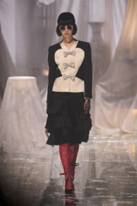 Model on a runway wearing a cream bodice with three black bows, black sleeves, and a black tiered skirt, paired with red lace tights, black shoes, round sunglasses, and a black hat.