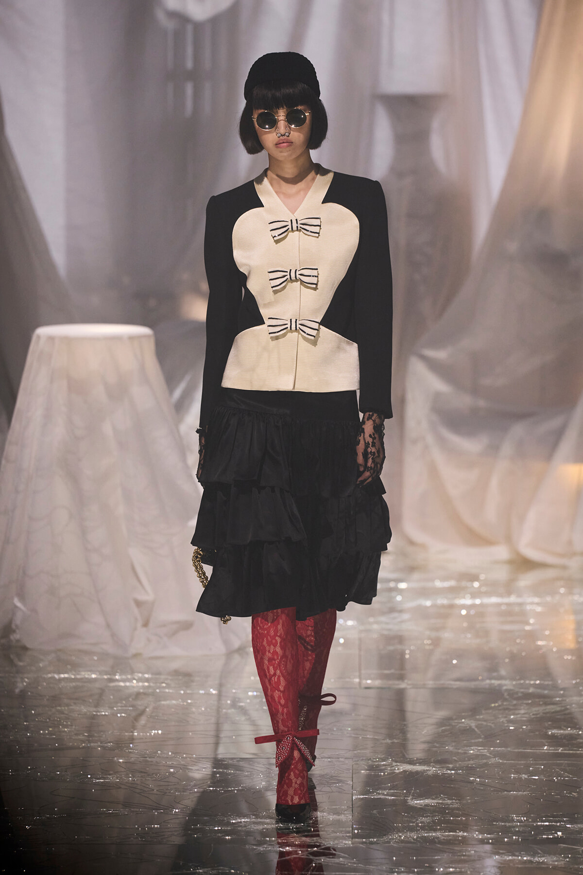 Model on a runway wearing a cream bodice with three black bows, black sleeves, and a black tiered skirt, paired with red lace tights, black shoes, round sunglasses, and a black hat.