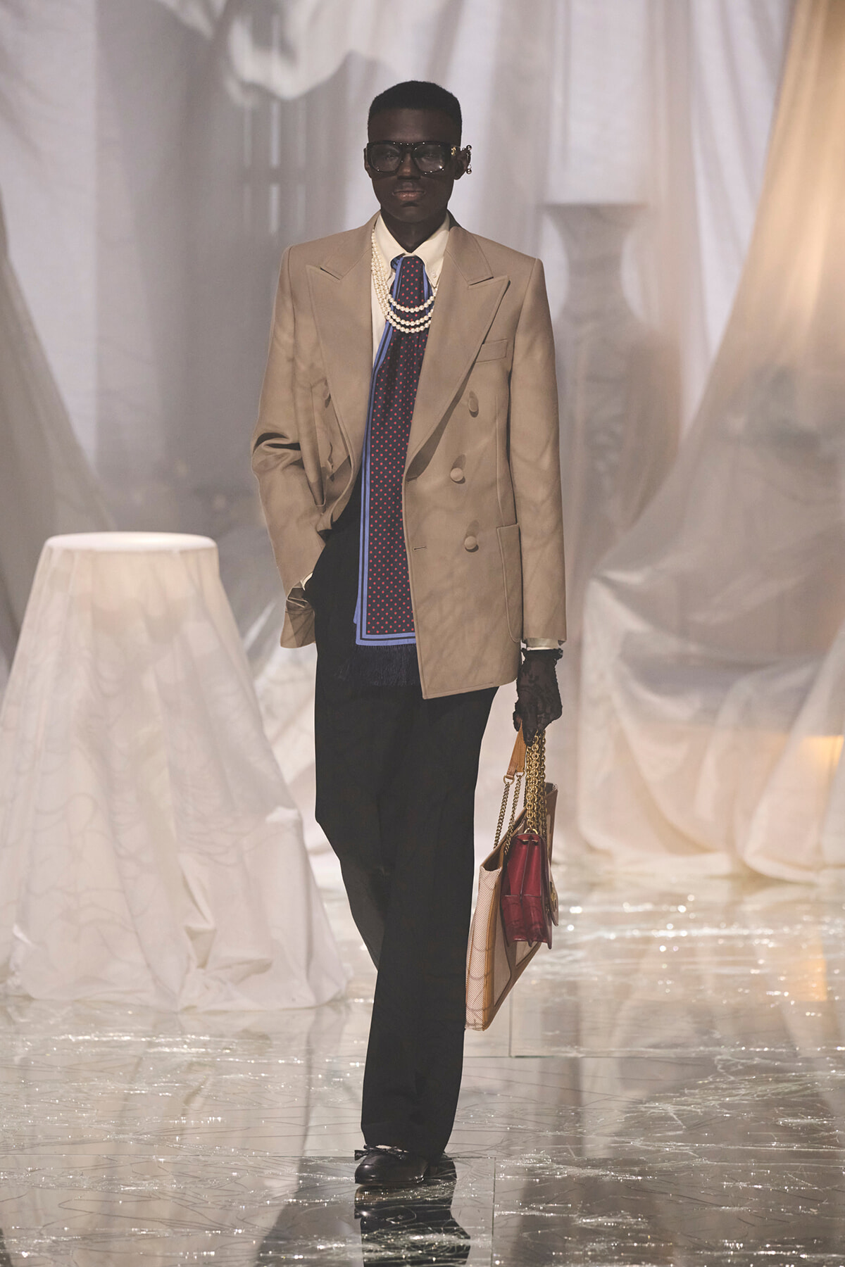 Runway model in a beige double-breasted blazer with pearl necklaces, a blue dotted tie, black trousers, gloves, and a beige handbag.