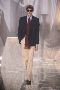 Runway model wears an oversized navy blazer, red polka-dot scarf, beige wide-leg trousers, and dark sunglasses, with a burgundy knit cap and patterned glove.