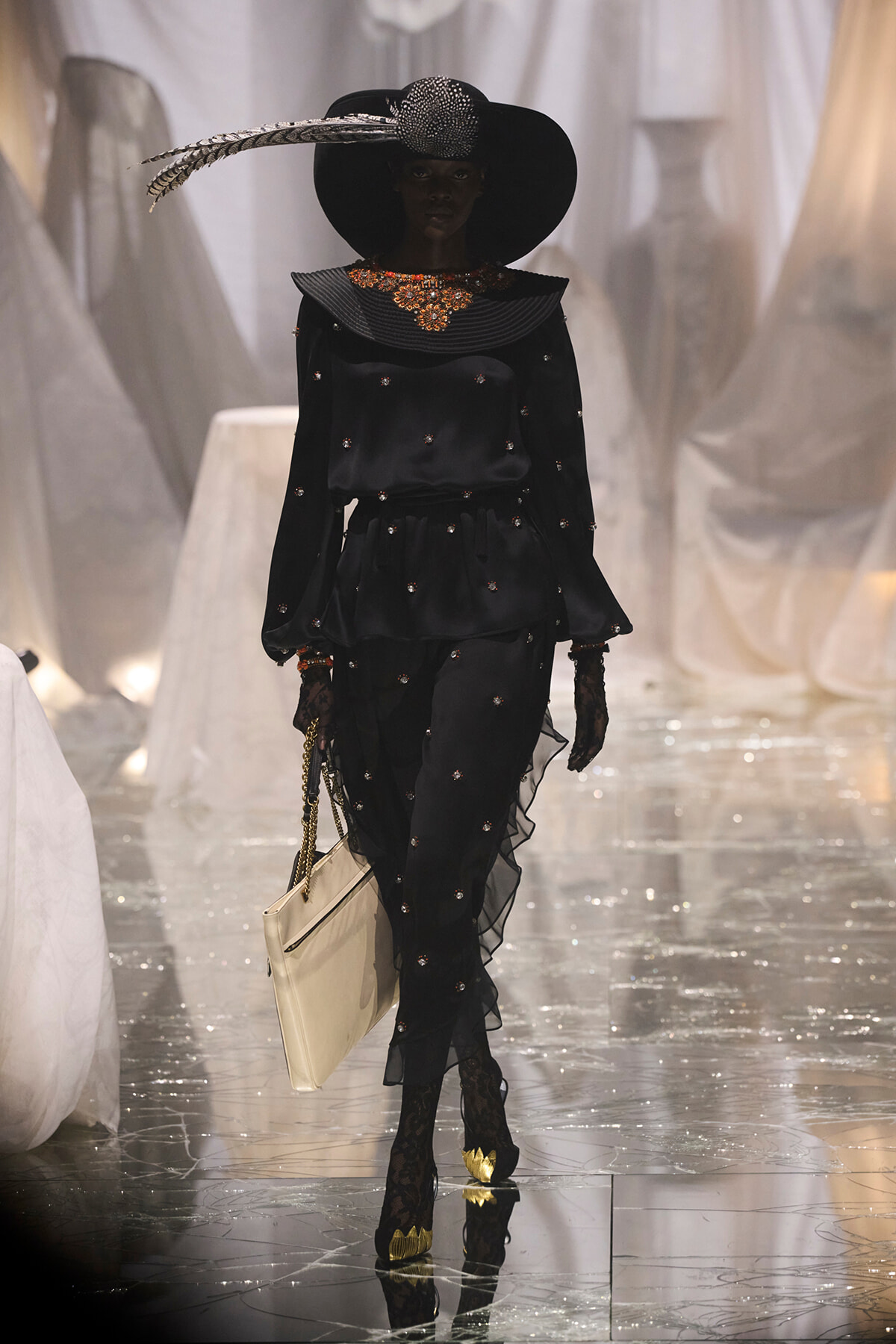 Model wearing a dramatic black ensemble with a wide-brim hat and feather, ornate neckline, and sheer gloves on a glossy runway.