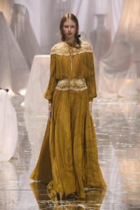 Model walking a fashion show in a mustard yellow, long-sleeve dress with white lace yoke and belt on a glossy runway.
