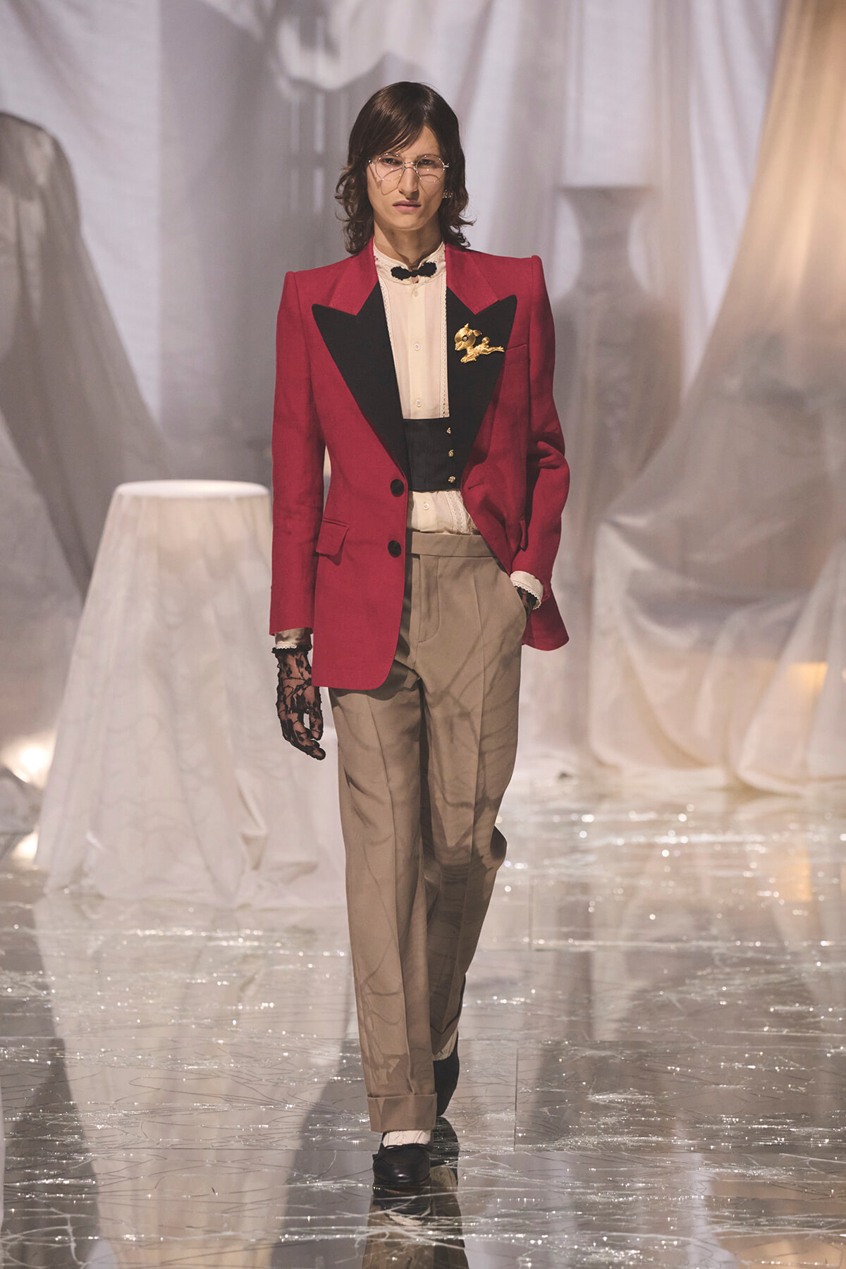 Model walking a runway in a red blazer with black satin lapels, cream shirt, beige trousers, and a black lace glove on one hand.