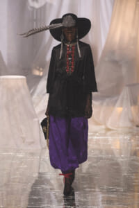 Runway model wearing a black wide-brim hat with a feather, a black embroidered tunic, purple puffed pants, and lace gloves while walking the catwalk.