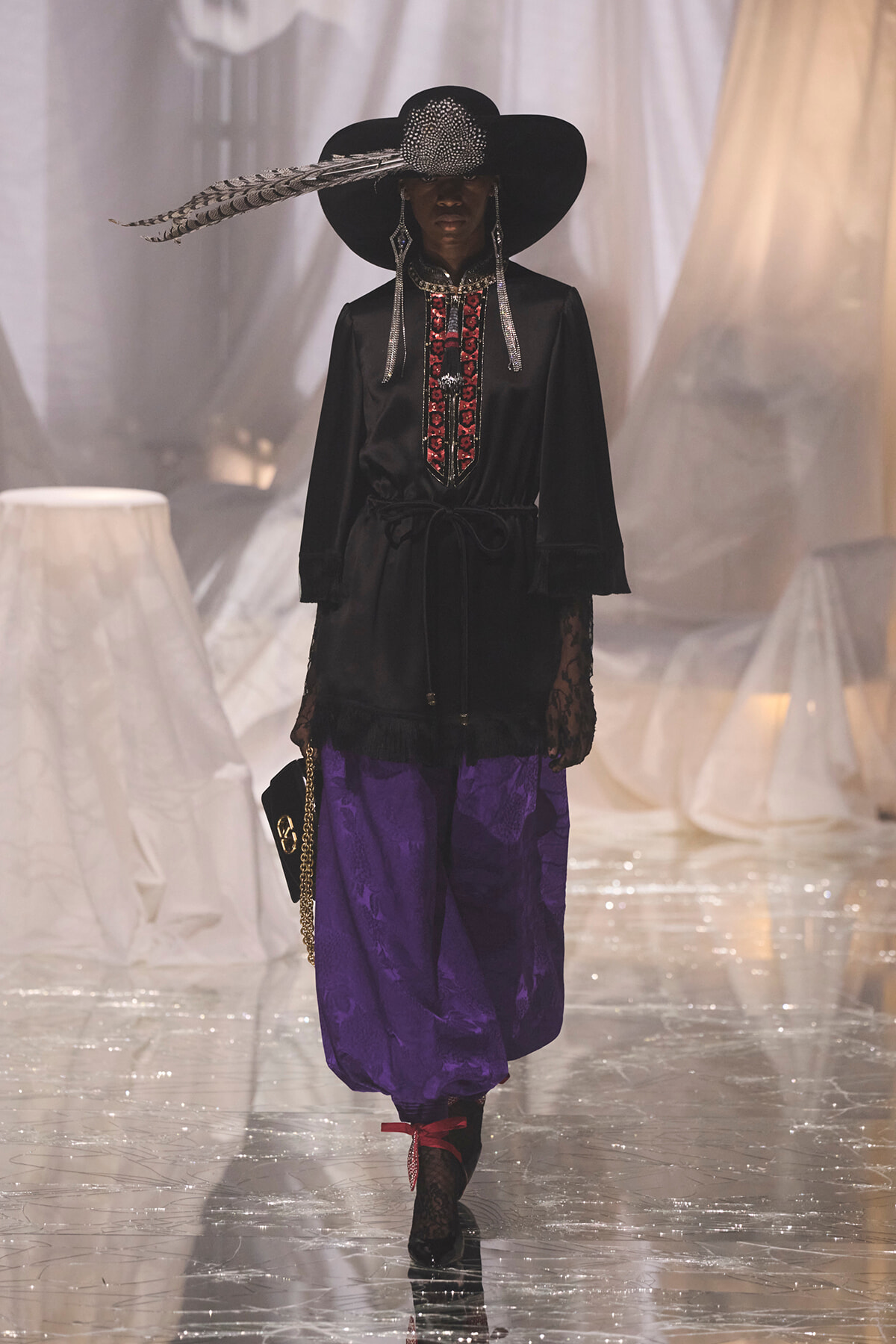 Runway model wearing a black wide-brim hat with a feather, a black embroidered tunic, purple puffed pants, and lace gloves while walking the catwalk.
