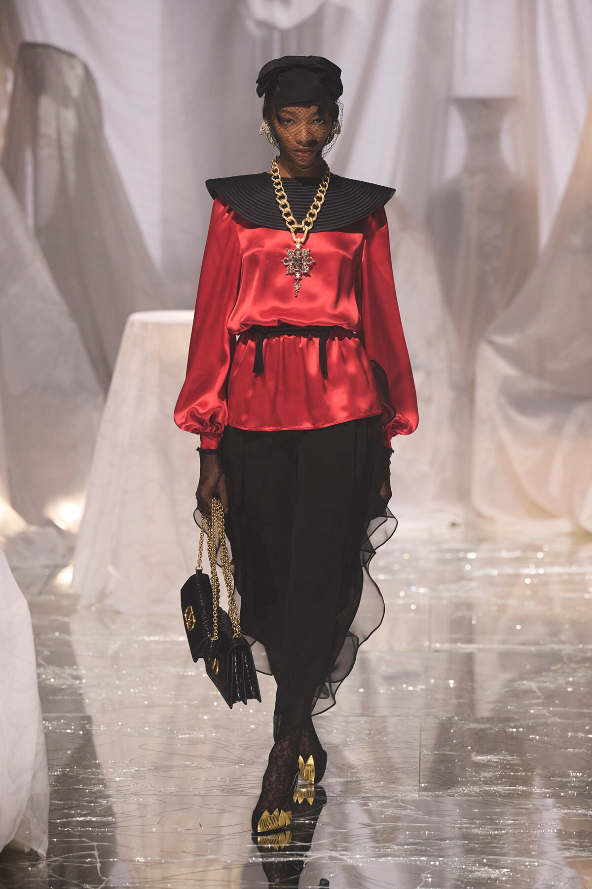 Model on a glossy runway wearing a red satin top with an oversized black collar, black sheer skirt, and gold jewelry while carrying a black purse with a chain.