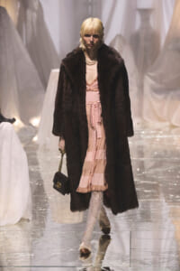 Model on a runway wearing a long dark fur coat over a pink dress with a belt, pearl necklace, and lace tights, carrying a small black handbag with a gold chain.
