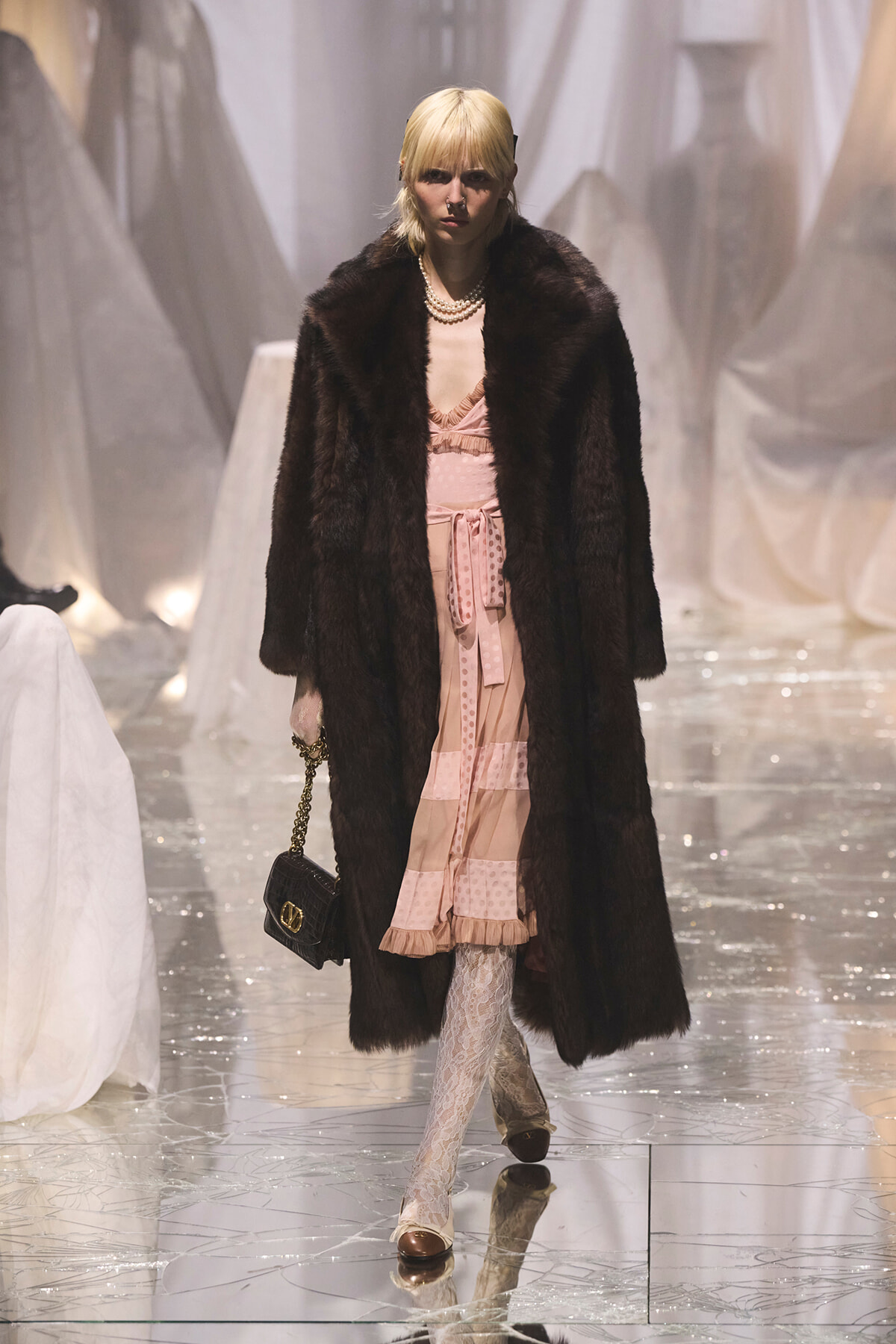 Model on a runway wearing a long dark fur coat over a pink dress with a belt, pearl necklace, and lace tights, carrying a small black handbag with a gold chain.
