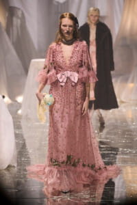 Model walking a fashion runway in a pink, beaded gown with a large satin bow at the waist; she holds a pastel doll accessory while another model in a fur coat appears blurred in the background.