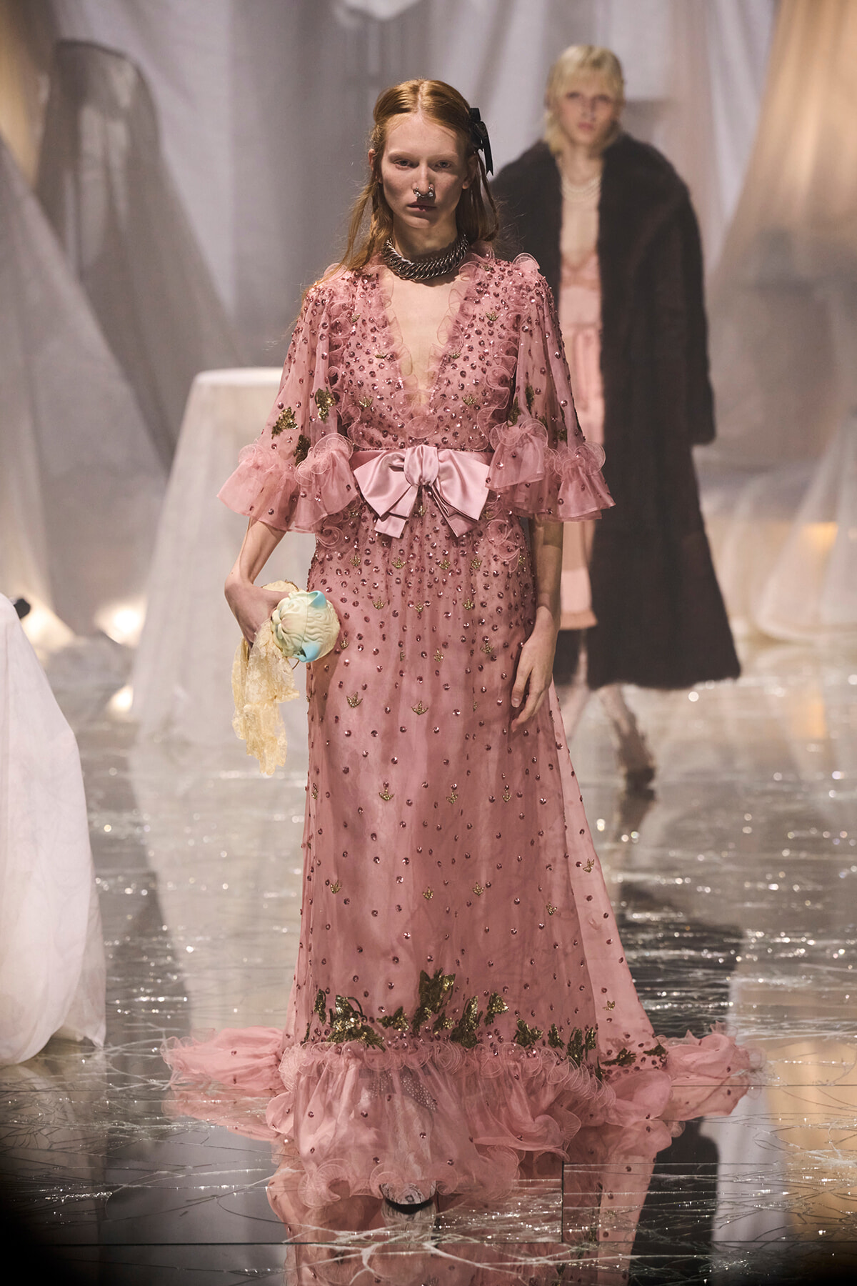 Model walking a fashion runway in a pink, beaded gown with a large satin bow at the waist; she holds a pastel doll accessory while another model in a fur coat appears blurred in the background.