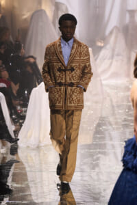 Male model walking a fashion runway in a brown patterned cardigan with toggle closures over a light blue shirt and tan trousers.