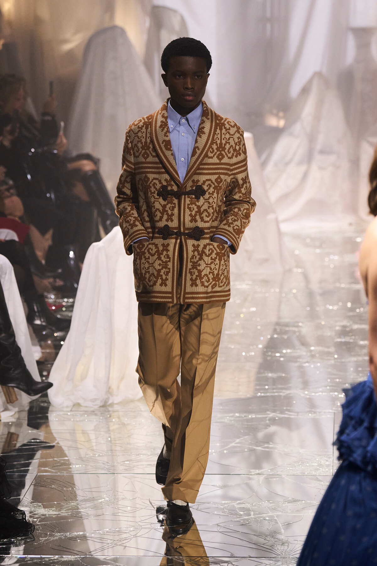Male model walking a fashion runway in a brown patterned cardigan with toggle closures over a light blue shirt and tan trousers.