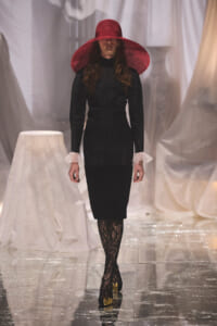Model in a black knee-length dress with sheer white cuffs and lace tights, wearing a wide red hat on a glossy runway.