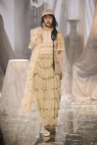 Model on a fashion runway wearing a cream Victorian-inspired lace gown with layered fringe and a fur stole on her shoulder.