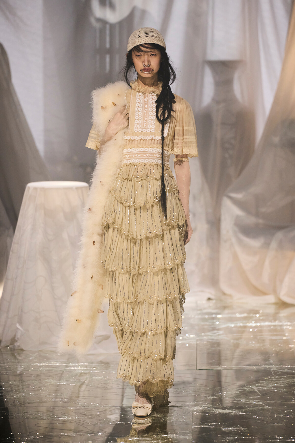 Model on a fashion runway wearing a cream Victorian-inspired lace gown with layered fringe and a fur stole on her shoulder.